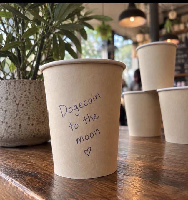 A brown paper coffee cup on a wooden counter has handwritten text reading 'Dogecoin to the moon' followed by a heart.