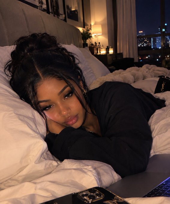 A woman with dark hair styled in a high bun lies on a bed with white pillows, wearing a black top and resting her chin on her hands, with a city skyline visible through the window behind her.