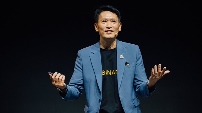 A man in a blue blazer with a 'BINANCE' branded t-shirt speaks on stage, gesturing with his hands against a dark background.