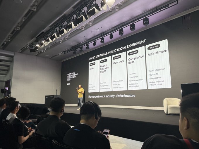 A presenter speaks on stage at what appears to be a conference, with a large screen behind them showing a slide titled 'CRYPTO STARTED AS A GREAT SOCIAL EXPERIMENT' and categorizing crypto into 'Geeks', 'ICO + DeFi', 'Compliance Build', and 'Mainstream' eras.