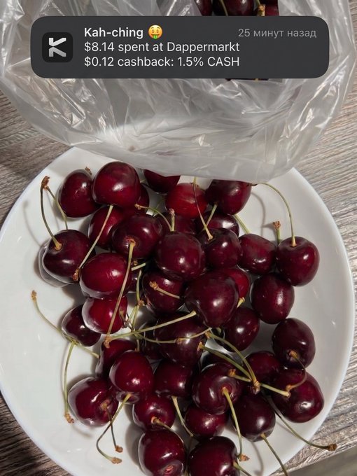 A white bowl filled with fresh red cherries sits beneath a partially visible plastic bag, with a notification overlay showing "Kah-ching 😋 $8.14 spent at Dappermarkt, $0.12 cashback: 1.5% CASH." The image illustrates the satisfying crypto-powered impulse purchase described in the post, where a quick craving was satisfied and paid for seamlessly using a KAST card.