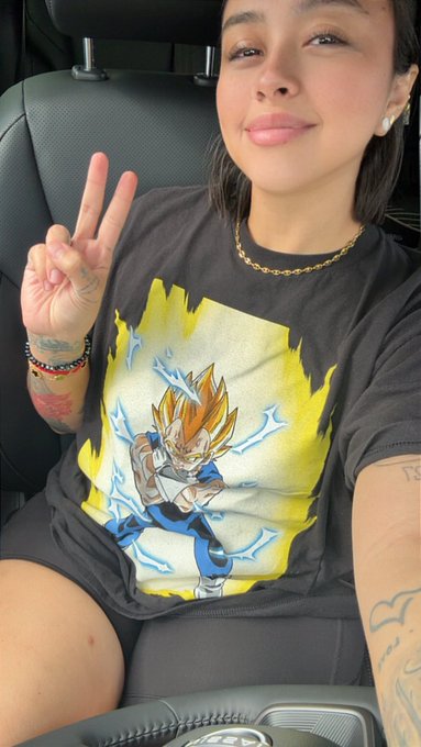 A woman in a car makes a peace sign while wearing a Vegeta (Super Saiyan) graphic t-shirt, gold chain, and pearl earrings.
