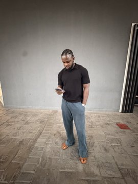 A man in a black polo shirt and jeans stands barefoot on a tiled floor, looking down at his phone with a smile—likely checking the "TP hit" (take profit) he mentioned in his post celebrating trading gains and inviting others to "leave Gold" for him.