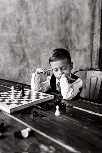 The image shows a young boy sitting at a table with a chessboard in front of him. He appears to be playing a game of chess, as there are two pieces on the board - one white piece and one black piece. The boy is wearing a suit and tie, which adds an air of formality to his attire while he plays. The image also contains text that reads "Happy New Week this bear market is bearing me sadly the quantum opportunity is in building the network that makes quantum compute accessible, verifiable and economically useful @QuipNetwork is creating the world's shared quantum computer". This suggests a connection between the boy's chess game and the development of new technologies like the Quantum Computer.