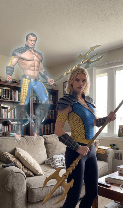 A woman in blue and yellow Aquaman-themed armor holding a trident stands in a living room, with a muscular Aquaman figure appearing to float or hover in the background.