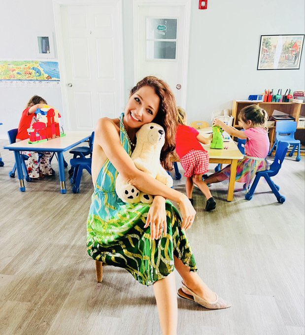 A smiling woman in a green and blue tropical dress holds a stuffed polar bear toy while sitting in a classroom with children playing in the background.