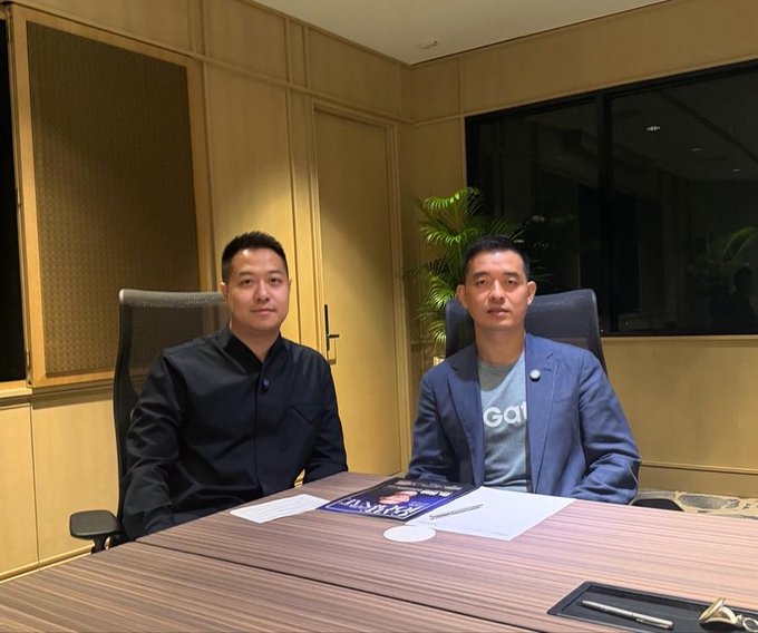 Two men sit at a conference table in a warmly lit office, with the man on the right wearing a blue blazer over a gray "Gate" branded t-shirt, accompanied by a magazine or document bearing the Gate logo on the table. The image captures the interview moment referenced in the post, featuring Han, CEO of @Gate, discussing his crypto journey since 2013 alongside the host.