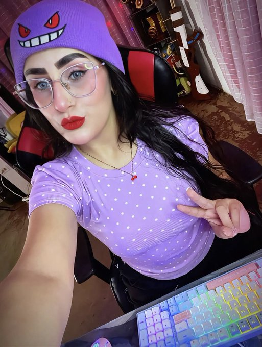 A selfie of a young woman wearing a purple Gengar beanie and matching polka-dot top, posing with a peace sign in front of a colorful gaming keyboard.