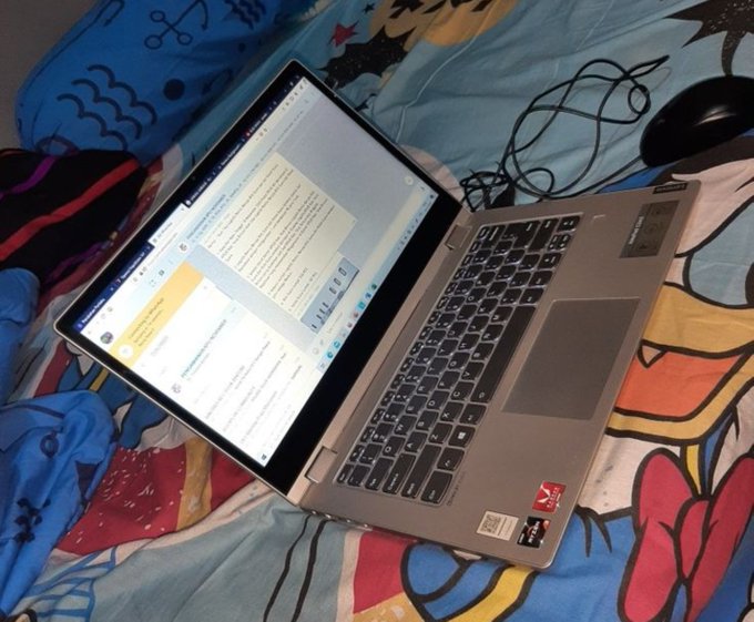 A silver laptop sits open on a colorful patterned bedspread, displaying a document with text on its screen, with a black computer mouse and cables nearby.