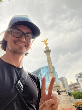 A selfie of a smiling man with glasses and a cap making a peace sign, standing in front of a tall monument with a golden winged figure on top, with modern city buildings in the background.