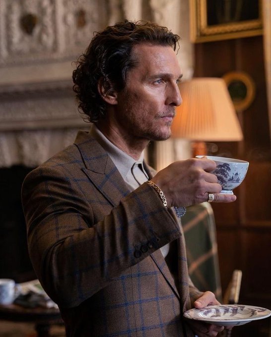 A man with curly hair, wearing a brown plaid jacket, holds a teacup and saucer in an elegantly furnished room.