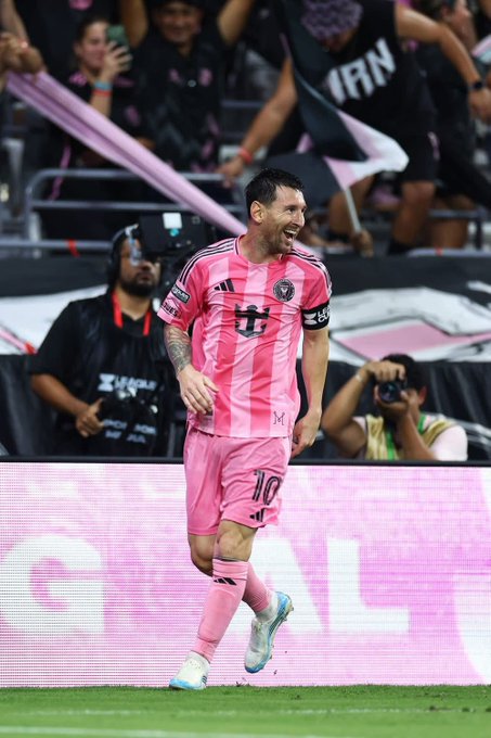 Lionel Messi, wearing a pink Inter Miami jersey with the number 10, celebrates on the field while fans wave banners in the background.