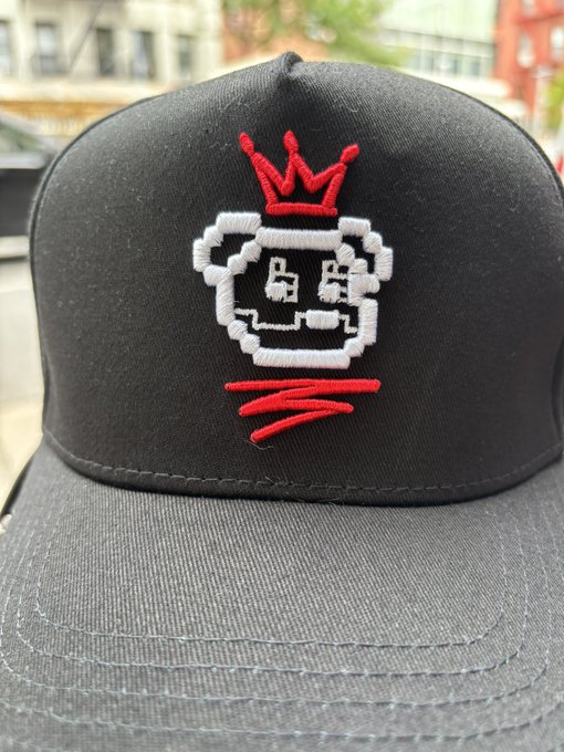 A close-up of a black baseball cap featuring an embroidered logo of a pixelated character wearing a red crown, with red scribbled lines beneath.