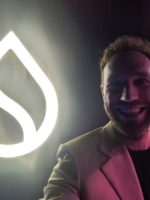 A smiling bearded man in a tan blazer poses next to a glowing white logo resembling a stylized 'S' or leaf shape.