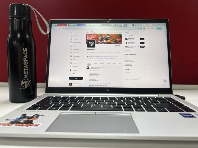 A laptop displaying the X (formerly Twitter) social media platform is shown on a desk next to a black Metaspace-branded water bottle, with the username 'MetaspaceX' visible on the screen.