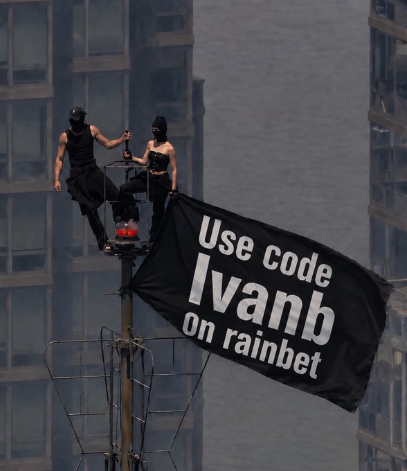 Two masked figures in black perch on a tall pole atop a city rooftop, unfurling a large black flag that reads "Use code Ivanb on rainbet," promoting a Rainbet gambling giveaway with promo code IVANB offering leaderboards, lossback, and wager milestone rewards.