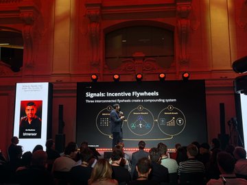 The image shows a man standing on stage giving a presentation about flywheels and their role as an open market for intelligence. There are several people sitting in chairs around him, attentively listening to his speech. The setting appears to be a large room with a stage at its center, where the speaker is delivering his talk. In the background, there are two TVs mounted on the wall, possibly displaying related content or visuals to support the presentation.