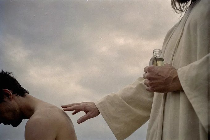 A robed figure holding a glass bottle extends a hand over the bare shoulder of a kneeling, head-bowed man beneath a cloudy sky, evoking a sacred anointing or baptismal scene that mirrors the post's theme of spiritual intention-setting and morning reflection.