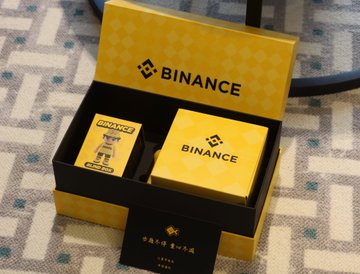 The image features a small toy robot sitting inside a box, which has the word "binance" written on it. The box also contains two other smaller boxes, one of which is yellow and the other black. These boxes are placed next to each other in the foreground of the image. The background of the photo shows a gray carpeted floor with a chair visible behind the robot toy.