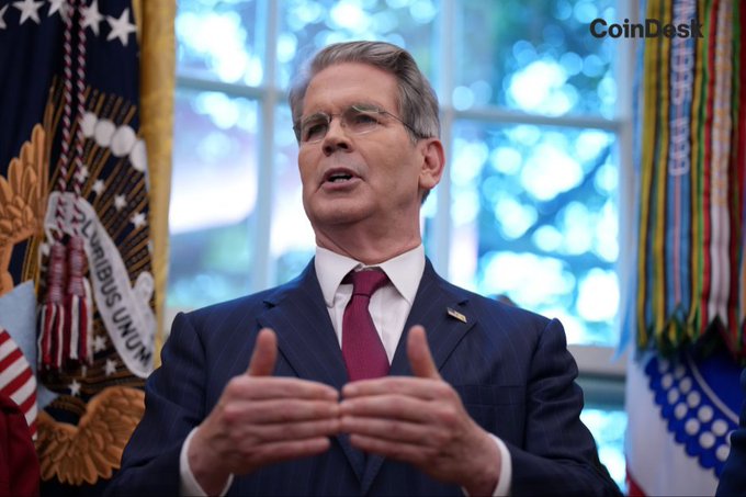 A man in a suit and red tie stands at a podium between flags, including the U.S. presidential flag, gesturing with his hands while speaking, with a 'CoinDesk' watermark visible.