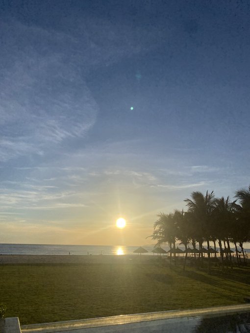 The sun rises over a serene beach lined with palm trees and thatched umbrellas, casting a warm golden glow across the calm sea and green lawn, perfectly symbolizing the hopeful start of a new week.