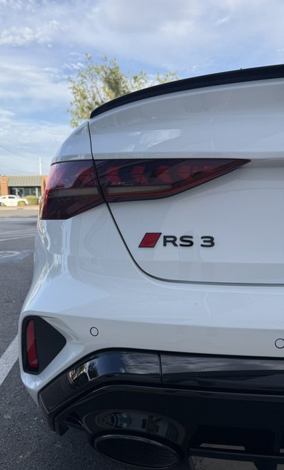 A close-up rear view of a white Audi RS3 sedan showcasing its "RS3" badge, dual oval exhaust tips, and integrated rear spoiler, presented as a proud new daily driver.