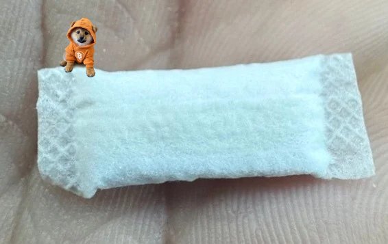 A tiny figurine of a small dog in an orange hooded outfit sitting on a white textured fabric or pad, placed on a person's palm.