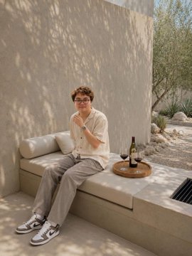 A man with glasses sitting on a tan outdoor cushioned bench holding a glass of wine, with a bottle resting on a nearby wooden tray.