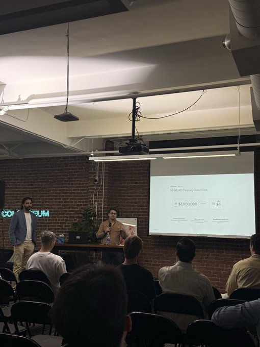 Two presenters at the MetaDAO event in front of a brick wall, with a slide showing 'MooDAO Treasury Conversion' figures of $2,000,000 and 3.56, while an audience watches.