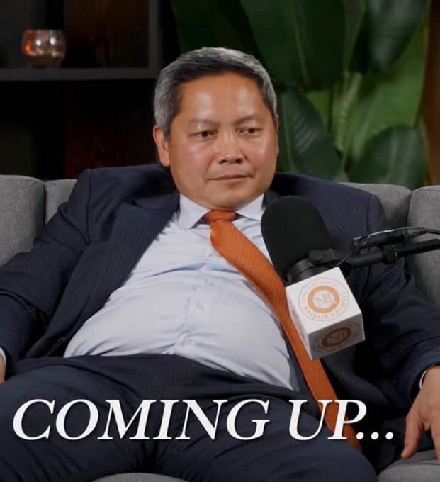 A man in a navy suit and orange tie reclines in a chair with a microphone in front of him, with text overlay reading "COMING UP..."—a humorous look at a relaxed executive that the post jokingly cites as a reason to buy MSTR.