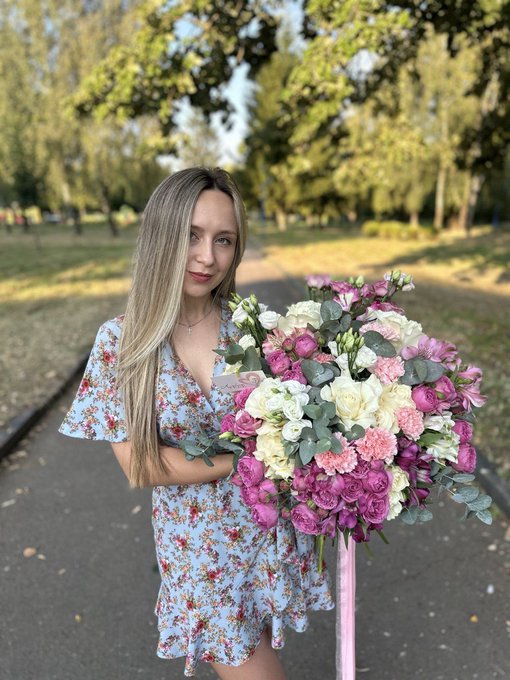 A young woman with long blonde hair wearing a floral dress stands in a park holding a large bouquet of pink, white, and cream-colored flowers including roses and lisianthus.