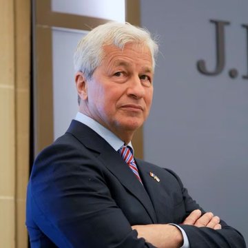A portrait of an older man with white hair wearing a dark suit and American flag tie, identified by context as Jamie Dimon, CEO of JPMorgan Chase, photographed in front of a J.P. Morgan branded background.