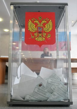 A transparent ballot box adorned with the Russian coat of arms is filled with folded paper ballots, depicting the ongoing parliamentary election mentioned in the post about which party will secure the most seats, with bets favoring United Russia.