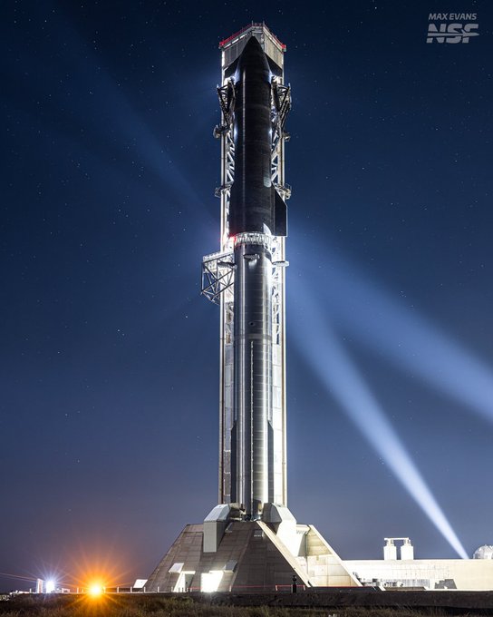 A nighttime photograph features a tall, fully stacked SpaceX Starship rocket illuminated by bright spotlights, with text crediting the photo to 'MAX EVANS NSF'.