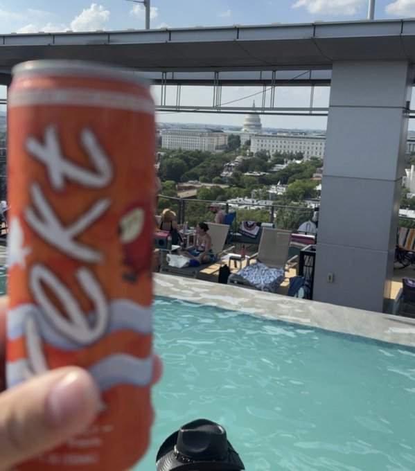 A hand holds an orange 'KOKO' branded soda can in the foreground while a rooftop pool offers a scenic view of the U.S. Capitol building in Washington, D.C.