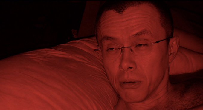 A close-up selfie of an older man wearing glasses, lying on a pillow in dim, reddish lighting.