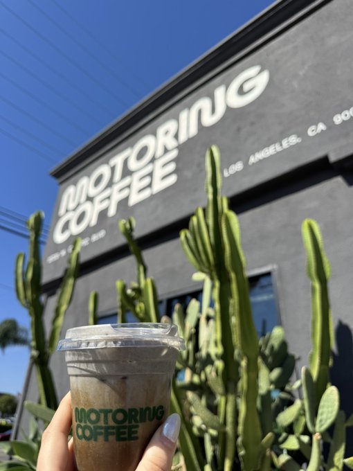 A hand holds a clear plastic cup of iced coffee with 'MOTORING COFFEE' printed in green, posed in front of the dark gray 'MOTORING COFFEE' building in Los Angeles, CA with cacti in the foreground.