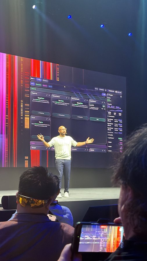 A man presents on stage at a tech conference, gesturing toward a large screen displaying a dashboard with tools like TrendSpot, BuzzPulse, and ScoreAI, while audience members watch and one films on a smartphone.