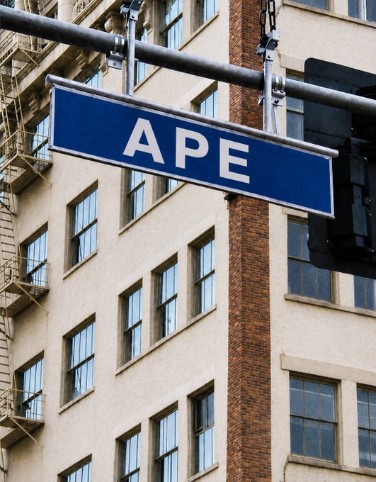 A blue street sign reading "APE" hangs from a metal pole in front of a tan brick building with fire escapes and large windows, cleverly evoking the $APE ticker mentioned in the accompanying post.