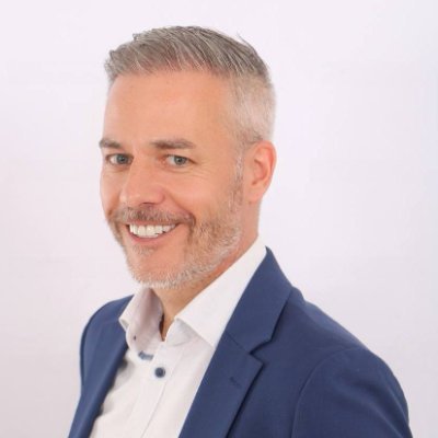 A professional headshot of a smiling middle-aged man with short gray hair and a neatly trimmed gray beard, wearing a navy blue blazer over a white collared shirt, posed against a plain white background.
