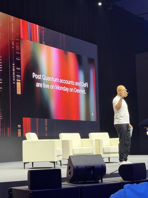 A man in a white shirt speaking on a stage with a large screen displaying 'Post Quantum accounts and DeFi are live on Monday on Devnet.'