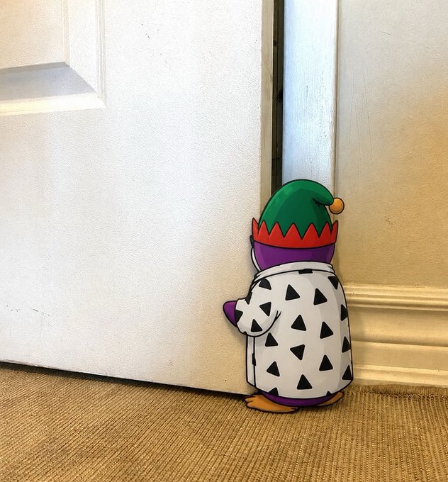 A wooden cutout decoration of a penguin wearing a green elf hat and patterned coat, tucked behind a slightly open white door.