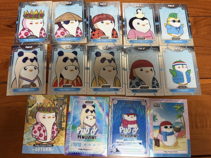 A collection of Pudgy Penguins trading cards and packs arranged on a wooden surface, featuring various penguin characters in different outfits and scenes.