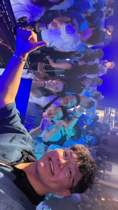 A young man in glasses smiles and flashes a thumbs-up in the foreground of a crowded club or event venue bathed in blue lighting, with a lively group of friends posing behind him.

The celebratory group photo reflects the post's sentiment of sharing a competition victory with friends, with the man proudly acknowledging their team effort despite not winning first place, promising to split the prize next time.