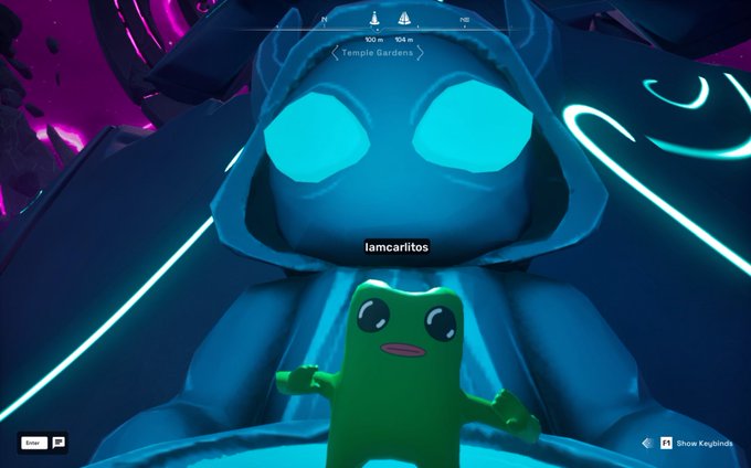 A green amphibian-like creature from the Koda collection in OthersideMeta is posed in front of a large blue robotic figure with glowing cyan eyes, with the username "lamcariltos" displayed and a location marker indicating "Temple Gardens" at coordinates 100m and 104m. The image aligns with the post celebrating the user @itsafwog receiving their #KODA companion in the OthersideMeta virtual world.