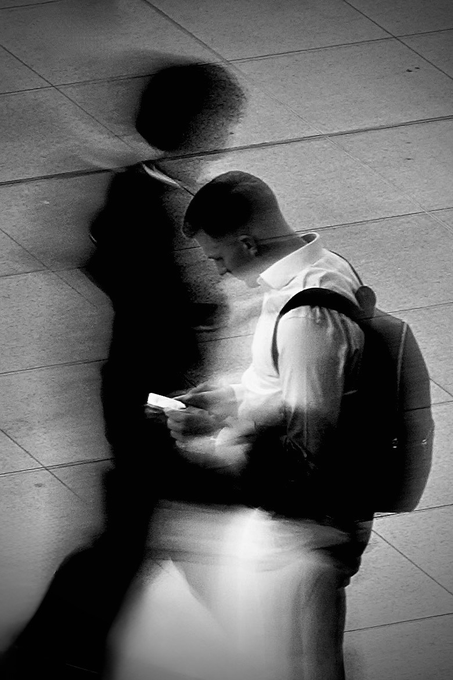 A black and white photograph with motion blur showing a person in a cap and backpack looking at their phone, with a distorted shadow cast against a tiled wall.
