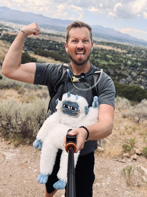 A man flexes his arm while holding a selfie stick and a white yeti plush toy, with a scenic valley and mountains in the background.