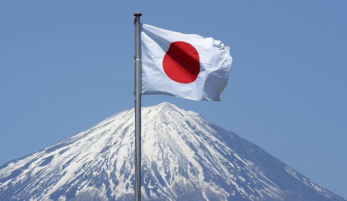 A Japanese flag waves against a clear blue sky with a snow-capped Mount Fuji visible in the background.