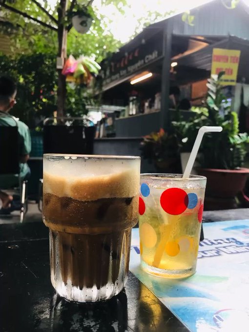 A glass of iced Vietnamese coffee with a thick layer of cream sits next to a colorful polka-dot glass of iced tea with a straw on an outdoor café table.
