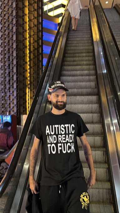 A bearded man wearing a cap with "I AM RETARDED" text and a black T-shirt reading "AUTISTIC AND READY TO FUCK" riding down an escalator in a modern building.